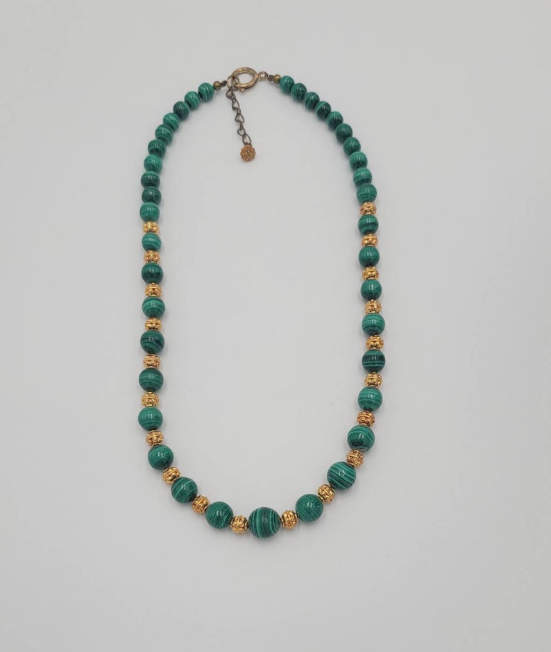 May include: A green malachite bead necklace with gold accents. The necklace is made of round beads and has a clasp closure.
