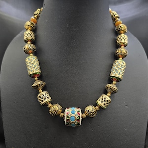 Könnte beinhalten: Eine goldfarbene Halskette mit einer Vielzahl von Perlen. Die Halskette weist zylindrische und kugelförmige Perlen mit komplizierten Mustern und farbigen Akzenten auf. Die zentrale Perle ist weiß mit blauen und roten Details. Die Halskette wird auf einer schwarzen Schaufensterpuppe präsentiert.