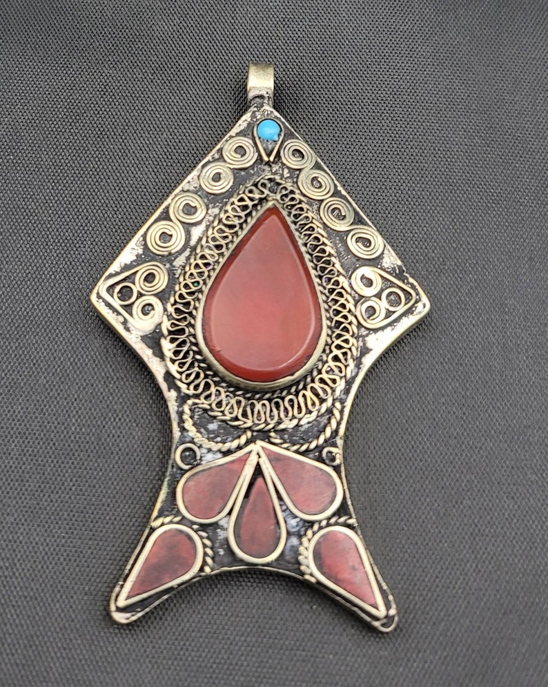 May include: A silver pendant with a red carnelian stone and a turquoise accent. The pendant is decorated with intricate wirework and has a fish-like shape.