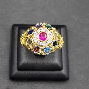 Puede incluir: Un anillo de oro con una gran piedra preciosa rosa rodeada de piedras preciosas más pequeñas de varios colores, incluyendo rojo, verde, azul y amarillo. El anillo está engastado en una banda de metal blanco.