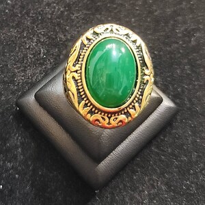 Könnte beinhalten: Ein goldener Ring mit einem ovalen grünen Edelstein, der in einem dekorativen Rahmen gefasst ist.