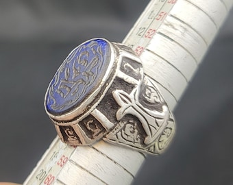 Può includere: Anello d'argento con pietra blu, caratterizzato da intagli intricati e un design dettagliato. L'anello è su un misuratore di anelli, che mostra le sue dimensioni e la sua maestria. La pietra ovale è incisa con un motivo ornato.