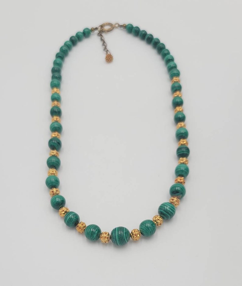 May include: A green malachite bead necklace with gold accents. The necklace is made of round beads and has a clasp closure.