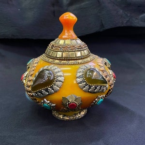 Op de afbeelding: Een gele keramische pot met een gouden deksel en decoratieve accenten. De pot is versierd met groene, rode en turquoise edelstenen.