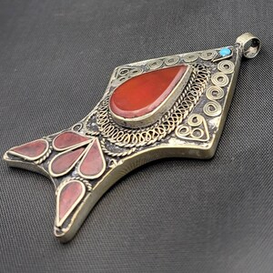 May include: A silver pendant with intricate filigree work and red gemstone accents. The pendant is shaped like a fish and has a small turquoise stone at the top.