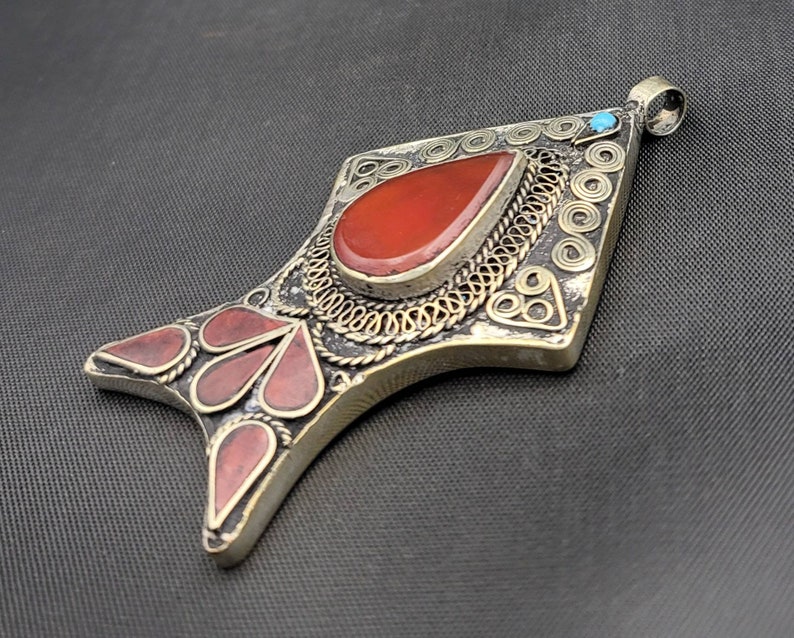 May include: A silver pendant with intricate filigree work and red gemstone accents. The pendant is shaped like a fish and has a small turquoise stone at the top.