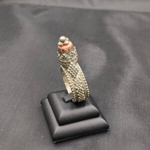 Può includere: Un anello d'argento con un designo testurizzato e una pietra di colore corallo sulla parte superiore. L'anello è esposto su un supporto nero.