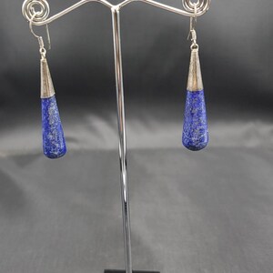 May include: A pair of silver dangle earrings with blue lapis lazuli stones. The earrings are hanging from a silver stand.