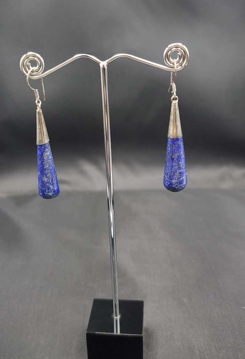 May include: A pair of silver dangle earrings with blue lapis lazuli stones. The earrings are hanging from a silver stand.