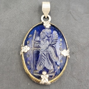 Peut inclure: Un pendentif en argent et or avec un camée en verre bleu représentant une figure d'ange sculptée. Le camée est entouré d'un cadre en argent avec des accents floraux décoratifs.
