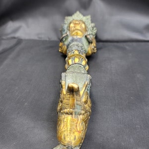 May include: A gold and green metal prayer wheel with a detailed design of a deity's face and a dragon-like figure.