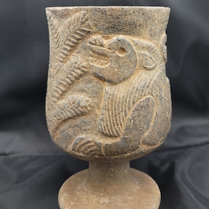 May include: A dark grey stone goblet with a carved design of a mythical creature. The goblet has a wide base and a rounded bowl.