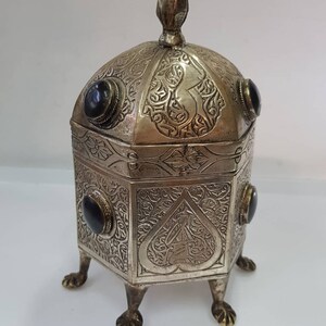 Puede incluir: Una caja de plata con intrincados diseños grabados y piedras preciosas negras. La caja tiene una tapa abovedada con un remate puntiagudo y se asienta sobre cuatro patas.