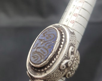 Può includere: Un anello d'argento con una pietra blu incastonata in una lunetta ovale. La pietra è incisa con disegni intricati. La fascia dell'anello è decorata con motivi floreali.