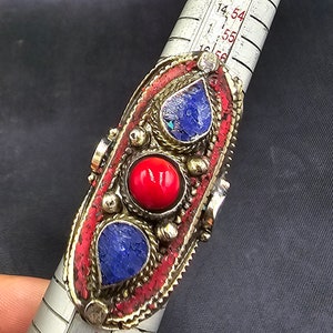 Könnte beinhalten: Ein silberner Ring mit roten und blauen Steinen, die in einem dekorativen Design eingefasst sind. Der Ring wird mit einem Lineal gemessen.