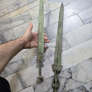May include: Two antique swords with ornate handles. The swords are made of metal and have a green patina. The swords are lying on a marble floor.