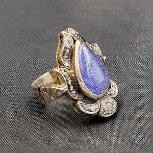 Op de afbeelding: Een zilveren ring met een traanvormige blauwe edelsteen omringd door kleine diamanten. De ring heeft een bloemmotief.