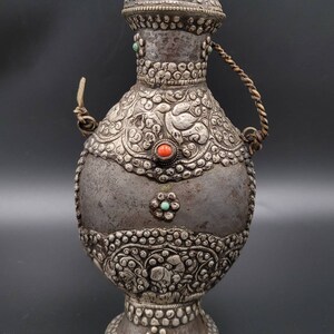 May include: An antique silver-plated water bottle with a round body, a small spout, and a rope handle. The bottle is decorated with intricate floral designs and has turquoise and red gemstones.