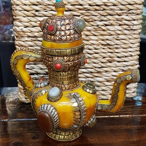 Op de afbeelding: Een gele theepot van keramiek met gouden en edelsteen accenten. De theepot heeft een rond lichaam en een tuit aan de zijkant. De deksel is versierd met een gouden band en een edelsteen.