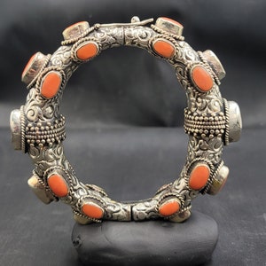 Puede incluir: Brazalete de plata adornado con piedras ovaladas de color coral. El brazalete presenta un detallado trabajo de volutas y se asegura con un cierre. Las piedras están espaciadas uniformemente alrededor de la circunferencia del brazalete.