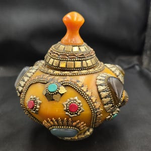 Op de afbeelding: Een sierlijke, amberkleurige pot met een deksel en een kleine oranje knop. De pot is versierd met goudkleurige metalen accenten, rode, turquoise en grijze stenen. De pot heeft een afgeronde vorm en is waarschijnlijk een decoratief item.