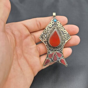 May include: A silver fish-shaped pendant with red and blue stones. The pendant is decorated with intricate wirework.
