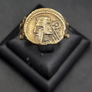 Könnte beinhalten: Ein goldener Ring mit einem detaillierten Design eines Personenkopfes auf der Oberseite. Der Ring befindet sich auf einem schwarzen Präsentationsständer.