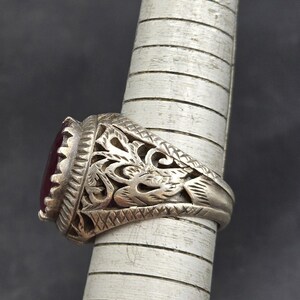 May include: A silver ring with an ornate design and a dark red gemstone. The ring is set on a finger-shaped display stand.