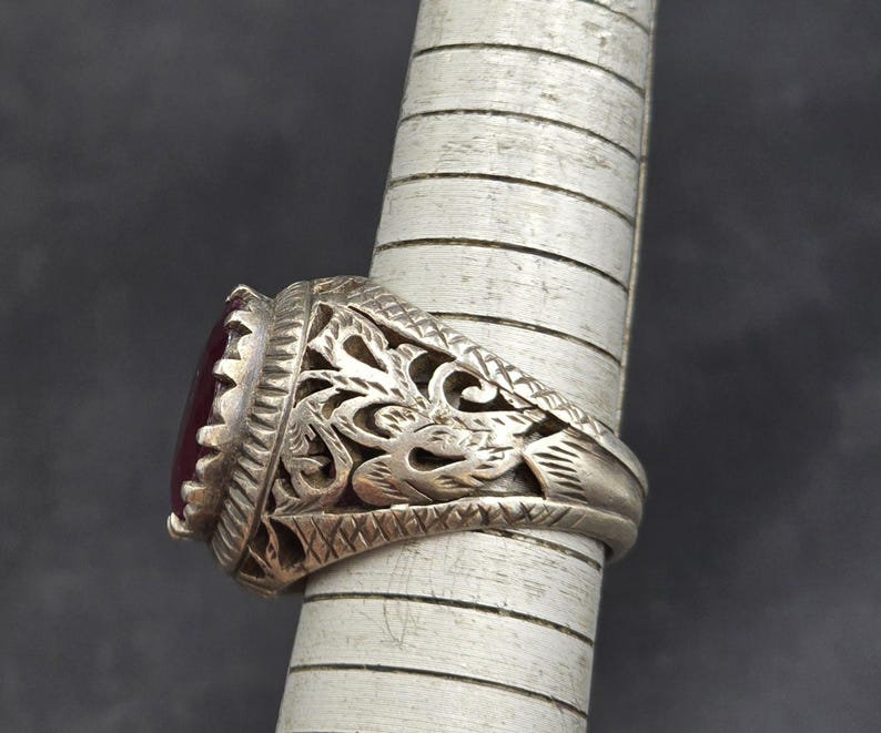 May include: A silver ring with an ornate design and a dark red gemstone. The ring is set on a finger-shaped display stand.