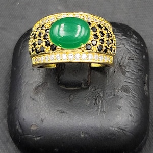 Könnte beinhalten: Ein goldener Ring mit einem großen, ovalen, grünen Edelstein, der von einem Band aus schwarzen und weißen Steinen umgeben ist.