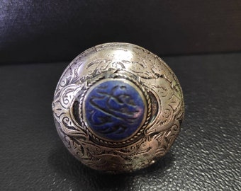 Può includere: Un anello d'argento con intricati disegni floreali e una pietra blu incastonata al centro. La pietra ha una scrittura araba incisa su di essa.