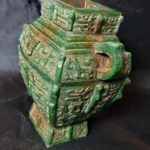 Puede incluir: Un jarrón de jade verde con tallas intrincadas y una base cuadrada. El jarrón está decorado con un patrón repetitivo de animales estilizados y diseños geométricos.