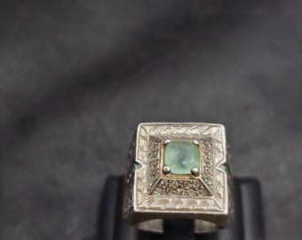 Puede incluir: Un anillo de plata con una piedra preciosa cuadrada de color verde esmeralda engastada en un marco cuadrado. El marco está decorado con grabados intrincados y pequeños diamantes.