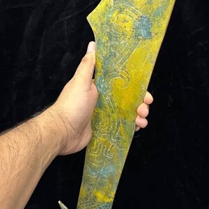 Puede incluir: Una espada grande de metal amarillo y verde con un mango de cabeza de dragón. La espada tiene tallas intrincadas y una hoja curva.
