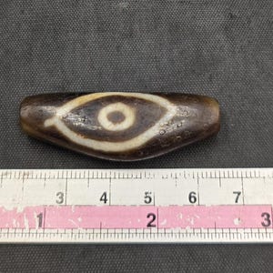 Puede incluir: Una cuenta de piedra cil&iacute;ndrica y alargada de color marr&oacute;n oscuro con un patr&oacute;n central en forma de ojo blanco y un centro circular marr&oacute;n oscuro. La cuenta est&aacute; colocada sobre una cinta m&eacute;trica blanca que muestra una longitud de 8,9 cm.
