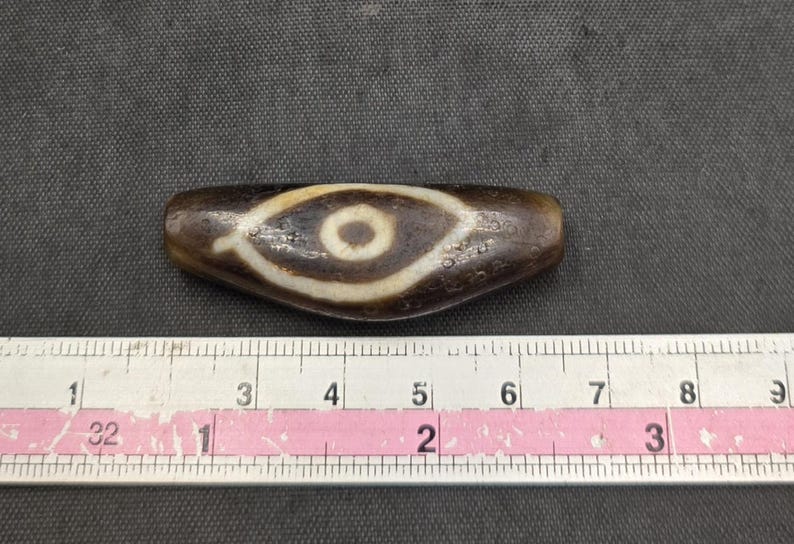 Puede incluir: Una cuenta de piedra cil&iacute;ndrica y alargada de color marr&oacute;n oscuro con un patr&oacute;n central en forma de ojo blanco y un centro circular marr&oacute;n oscuro. La cuenta est&aacute; colocada sobre una cinta m&eacute;trica blanca que muestra una longitud de 8,9 cm.