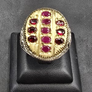 May include: A silver ring with a gold oval top set with red gemstones. The ring has a decorative border around the oval top.