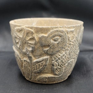 May include: A grey stone planter with a carved design of a stylised face and leaves.
