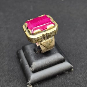 May include: A ring featuring a rectangular, vibrant magenta stone set in a textured, tarnished brass-toned metal band. The metal setting displays intricate geometric engravings and triangular patterns along the sides of the shank.
