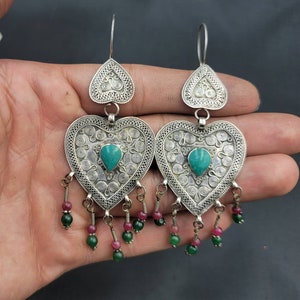 Op de afbeelding: Zilveren hartvormige oorbellen met turquoise stenen en groene en roze kralen die aan de onderkant hangen.