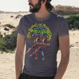 May include: A man wearing a grey t-shirt with a colourful graphic of a star and the text "The Brides of Guten Stein" in yellow. He is wearing a black baseball cap and standing in a desert landscape.