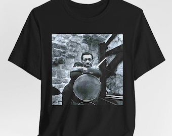 May include: A black short-sleeved t-shirt featuring a central square graphic print. The image displays a black and white photograph of a musician leaning over a drum, holding a drumstick, set against a stone wall and a dark wooden structure.