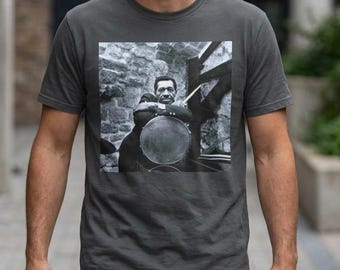 May include: A charcoal grey short-sleeved t-shirt featuring a square black and white photographic print on the chest. The graphic depicts a musician sitting behind a drum set, holding a drumstick, set against a stone wall and wooden structure.