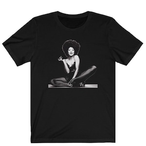 May include: Black and white photo of a woman with an afro sitting with her legs crossed. She is wearing a black leotard and black tights.