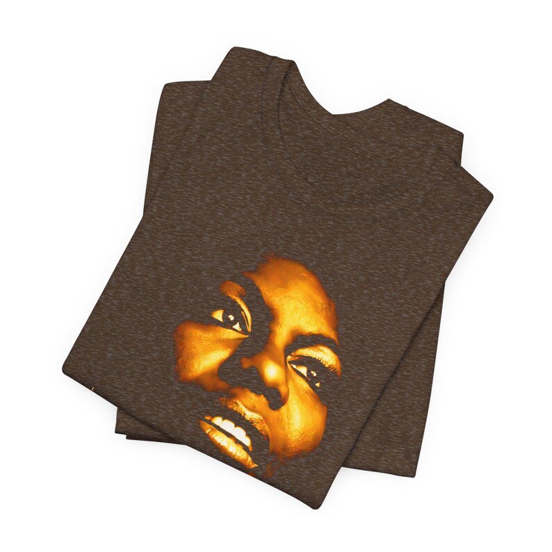 May include: A dark brown heathered t-shirt featuring a high-contrast, golden-yellow graphic print of a human face. The design focuses on the eyes, nose, and mouth, rendered in a warm, glowing tone that stands out against the textured fabric.