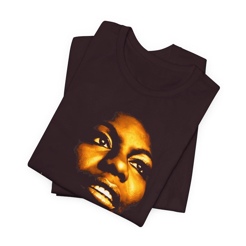 May include: A dark brown short-sleeved t-shirt featuring a high-contrast, golden-yellow graphic print of a person's face. The portrait focuses on the eyes, nose, and mouth, rendered in warm, glowing tones against the solid dark fabric background.