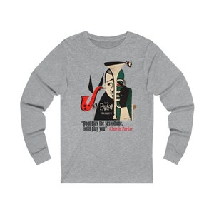 May include: A long-sleeve gray shirt featuring a stylized illustration of a saxophone player. The artwork includes a red saxophone, and the text "Charlie Parker Volume 2" and a quote by Charlie Parker.