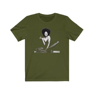 May include: A green t-shirt with a black and white image of a woman with an afro sitting with her legs crossed.
