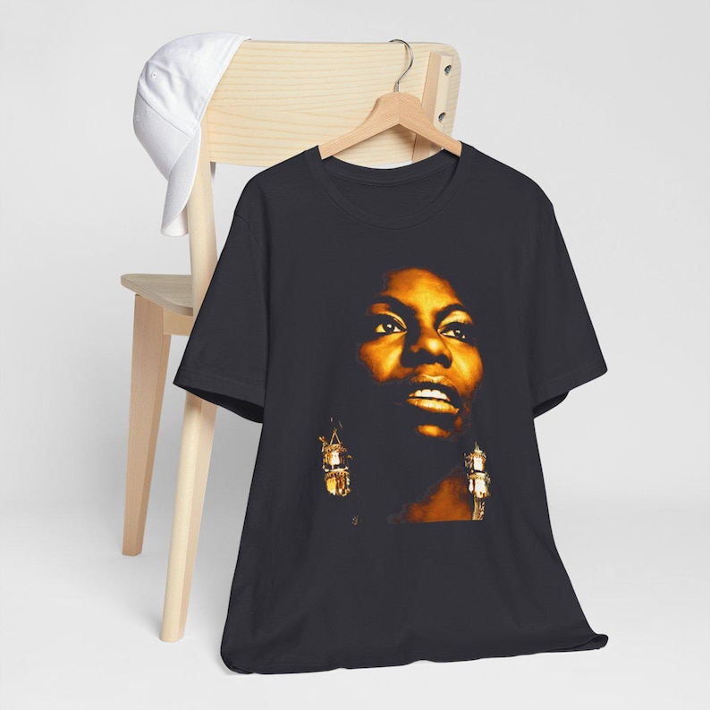 May include: A black short-sleeved t-shirt featuring a high-contrast, golden-toned portrait of Nina Simone on the front. The graphic depicts the singer with detailed facial features and prominent dangling earrings, set against the dark fabric background.