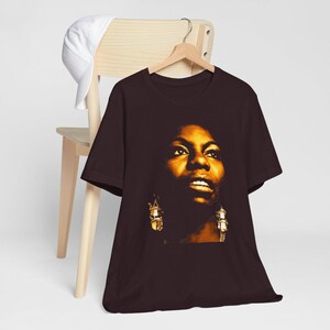 May include: A dark brown short-sleeved t-shirt featuring a high-contrast, golden-toned graphic portrait of Nina Simone. The print captures the face of the singer with detailed lighting, showing her wearing prominent dangling earrings.